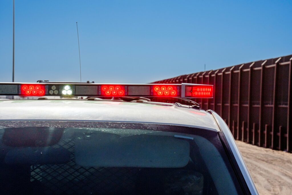 A close-up view of a police vehicle's roof with red and white emergency lights flashing, set against a bright blue sky and a long industrial fence running alongside a dusty road.