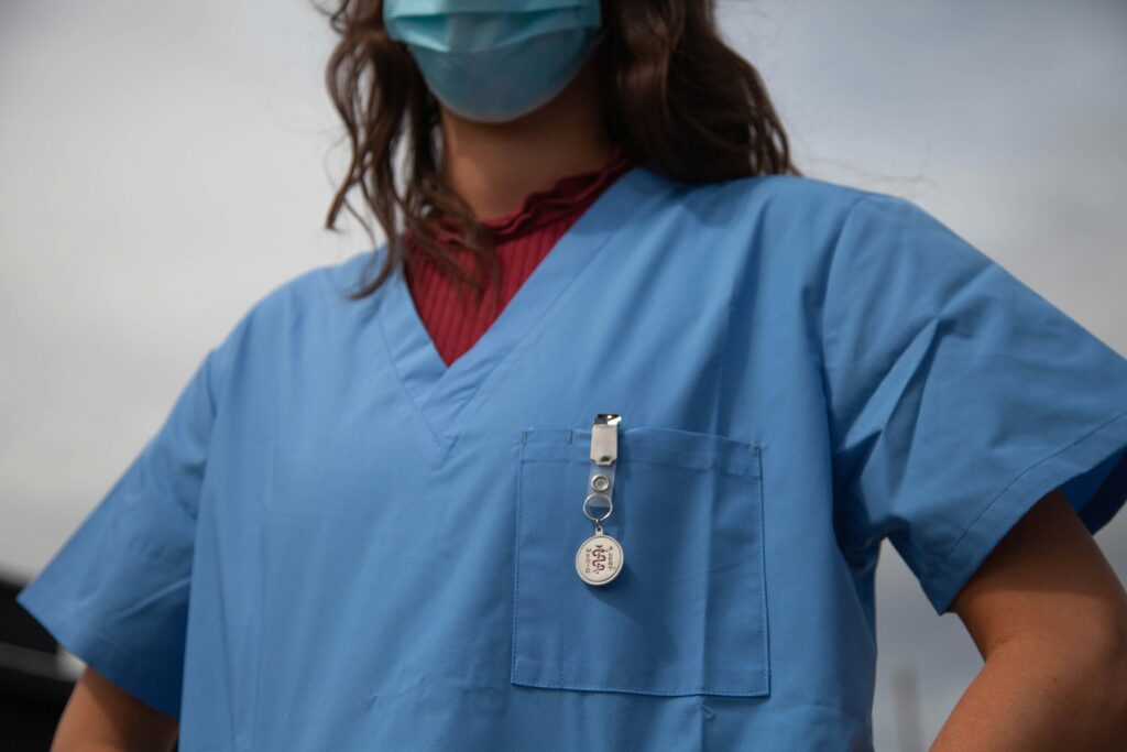 A healthcare worker wearing blue medical scrubs and a face mask stands with hands on hips, with a clipped ID badge visible on the chest pocket against an outdoor, overcast background.